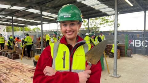 Tegan Pryor, in a high visibility vest, green helmet and holding a trowl