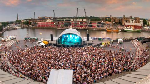 A performance at Bristol Sounds. The city's harbourside can be seen behind the stage, across the water. A large crowd are stood watching the performance. The stage is lit with blue lights. 