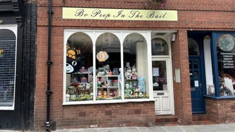 The outside of a shop window with three arched windows and a white door to the right. A black cat with an orange hat logo is peeking out from the left and there are several teddies on shelves in the window on display. At the top of the windows, a yellow and black sign reads "Bo Peep In The Bail".