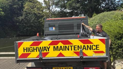 A man is putting a black bag of dead fish into the back of a Highway Maintenance van.
