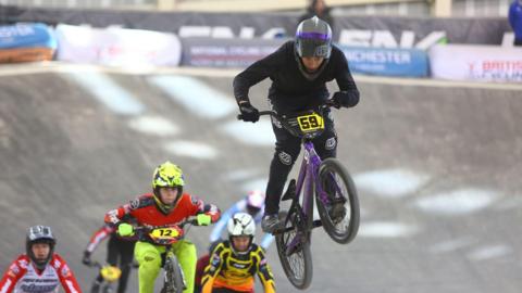 A teenage boy rides a BMX bike on a track. Other bikers can be seen behind him