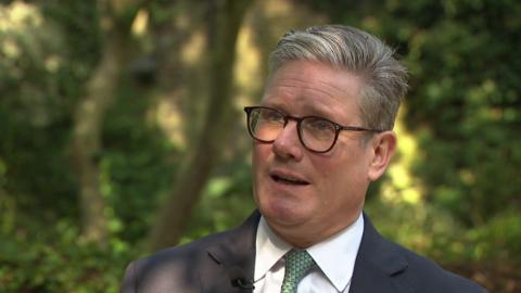 Sir Keir Starmer, wearing a dark suit, white shirt, tie and glasses, stands in a garden looking at the interviewer out of shot.