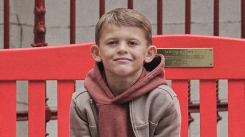 Archie has short brown hair and is wearing a pale red hoodie over a light brown jacket. He is smiling and sitting on a red bench.