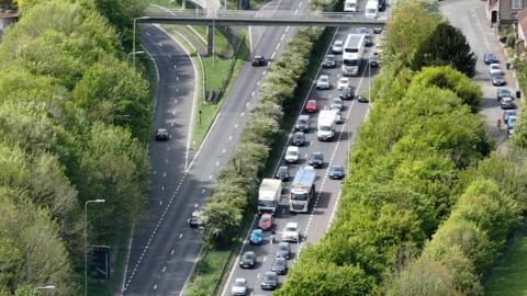 An aerial view of the A27 shows slow‑moving traffic queueing, with several lanes of cars and lorries backed up beside trees and a road bridge near a slip road.