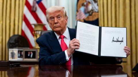 Trump holds up an executive order in the Oval Office, showing a pause in some tariffs on Mexico and Canada