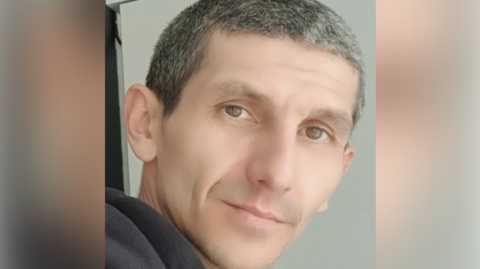 Man with short grey hair - he has brown eyes and is looking at the camera with his head turned to the right - he has a slight smile.