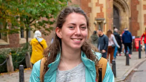 Gwennan yn sefyll o flaen coleg yng Nghaergrawnt. Mae hi'n gwisgo cot las ac yn dal beic.