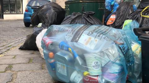 Rubbish in a blue clear bag on the kerbside. There are black bags around it with a green litter bin in the background.