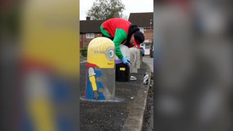 A bollard painted as a Minion, side view. A man behind it in red and green jacket is painting another one.