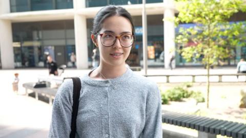 A woman with wide rimmed glasses, black hair and a grey knitted cardigan is looking to the left of the camera. In the background are trees and shops. 