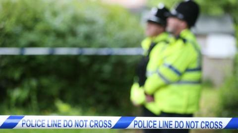 Two police officers in high vis jackets stand behind police tape