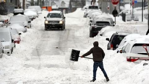 A man shovels out snow on a car lined road. Large amounts of snow surround the cars and a car is driving down the road.