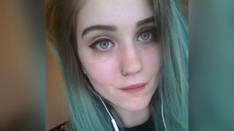 Tia Langdon from Southampton, a young woman with green/ blue hair, is looking at the camera and has a nose piercing and white headphones coming from her ears