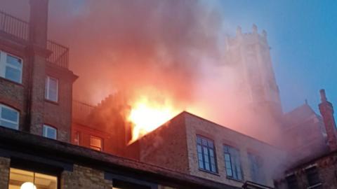 Flames and thick smoke rise from the upper floors of a brick building at dusk, with a church tower visible in the background.