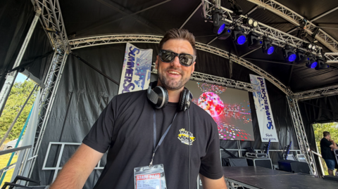 Ryan Swain, who is wearing a black shirt with a yellow and white logo that says You Me and ADHD, stands at a DJ mixing table with headphones around his neck. In the background is a large stage with a glitter ball that has been displayed on the screen behind him.