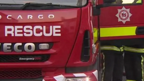 A red fire engine. One of the doors to the vehicle is open and there are two fire fighters in uniform standing by it.
