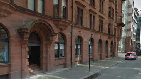 McClure Solicitor offices in Glasgow city centre, pictured in 2014. A red sandstone office building with arched windows and ornate masonry around the door has McClure signage in the windows. Modern office blocks are seen in the distance.