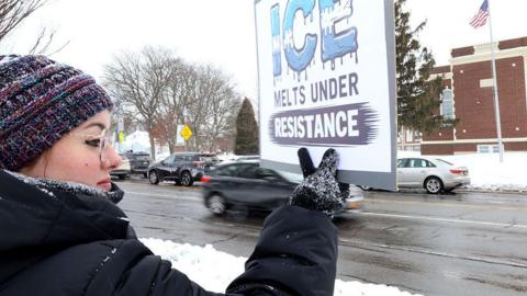 Protester, in profile, stands on a snowy roadside, wearing a tuque, glasses, and black winter coat and gloves, holds a sign that reads: "ICE melts under resistance."