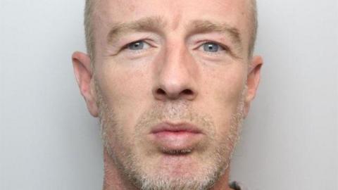 A mugshot of a middle-aged man with a a short grey beard and grey/blond  closely cropped hair. 