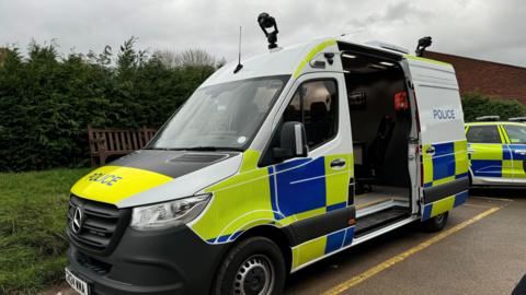 A van with blue, yellow and white police markings. It's middle door is open and two cameras are mounted to the top of it. It is parked next two another police car and a grass verge.