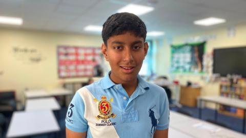 A boy in a blue polo shirt stands in the classroom of Manor High School 