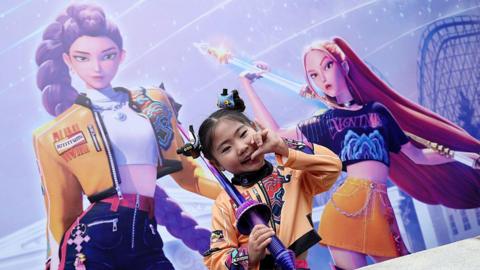 as little girl dressed as Rumi from Kpop demon hunters poses in front of a big poster of Rumi and Mira 