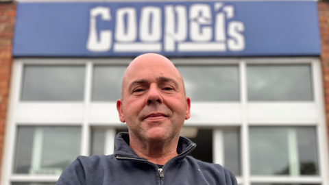 A head and shoulders image of Stuart Ruskin, taken from a low angle. He is looking into the camera and the Coopers logo is in the background above his head, blurred.