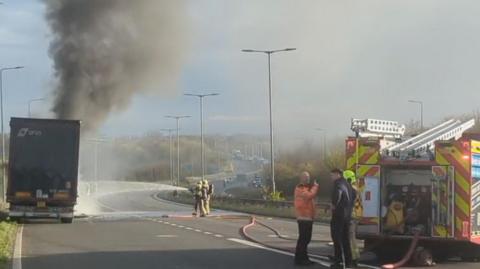 A fire crew uses a high-powered hose to dampen down a smoking lorry, which was on fire. There is a fire engine in the foreground.