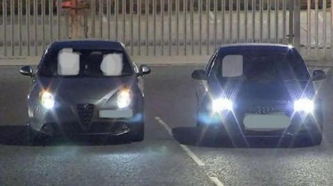 Two cars side by side with their lights on and reg plates blacked out, in what looks like an empty car park at night.
