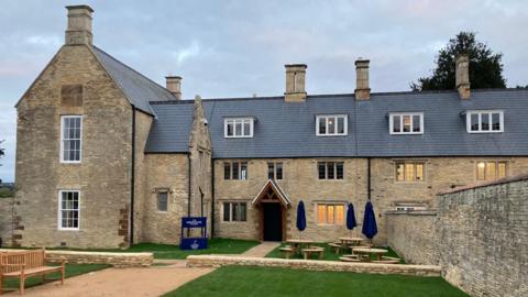 A large stone building with several windows and a dark slate roof. There is wooden entrance. In the front is small stone wall, with a larger wall to the right hand side. There is a also a green grass lawn and three picnic table. 