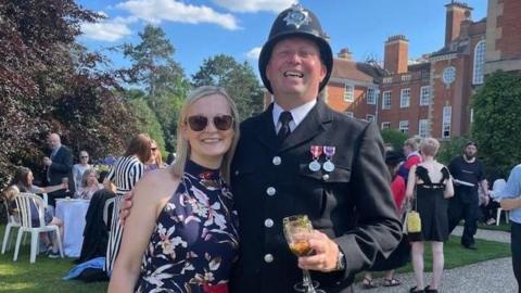 Police sergeant Laurence East in his uniform holding a glass of white wine in one hand with his other arm around his wife, Amy, who has blonde hair and is wearing a blue and white sleeveless dress.