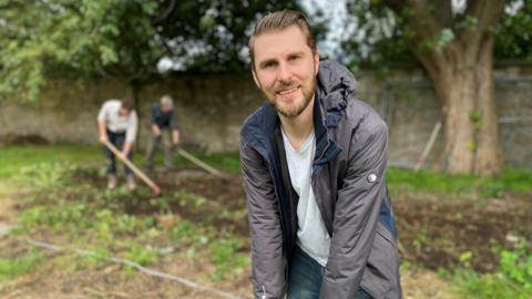 Scott Cambers, in a grey jacket, leans on a spade at the dig in a churchyard in Govan, with two other volunteers behind him combing through the soil