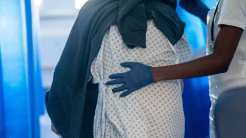 A person wearing a hospital gown with a small repeating square pattern is standing and being supported by a healthcare worker. The healthcare worker is wearing a white uniform and blue disposable gloves, with one hand placed gently on the patient’s back. A dark blanket or piece of clothing is draped over the patient’s shoulders. The background shows blue privacy curtains and a softly lit clinical setting.