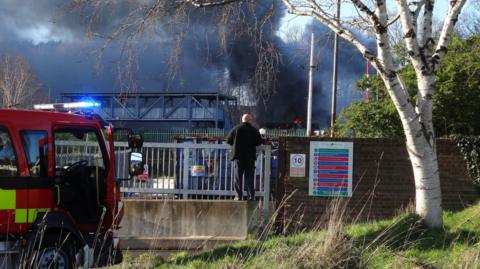 Plumes of black smoke are seen billowing from a building. There is a fire truck in front of the shot with a man standing on a platform looking over the railings at the fire