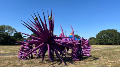 A puppet controlled by ten people which is bright purple with inflated spikes and four legs. It has a big spikey head and green adder eyes pointing out the top