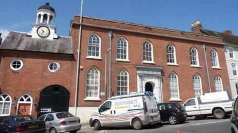 The Ludlow Guildhall building with vehicles parked in front of it. It is a two-storey building and a clock is on the left of the photo.