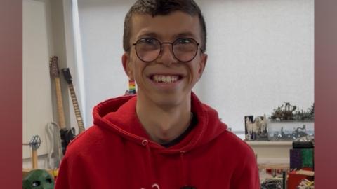 A young man wearing glasses smiles at the camera. He is wearing a red hoodie. In the background some guitars sit on a table. Arts and crafts are displayed on a windowsill.