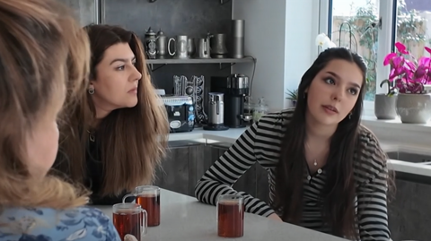 Two women sit at a kitchen counter with mugs of tea, one looking at the other, and the back of another woman's head as she sits facing them in the kitchen