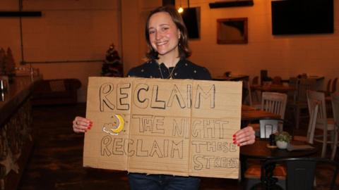 Woman with brown bob haircut smiles at camera, holding a sign which says 'Reclaim the Night, Reclaim These Streets'.