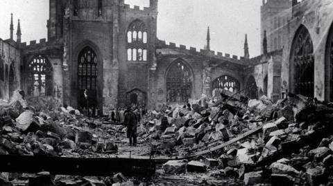 An old black and white photo showing inside a ruined cathedral with rubble strewn everywhere while the outer portion of the building remains standing. People can be seen entering the site and looking around at the scene.
