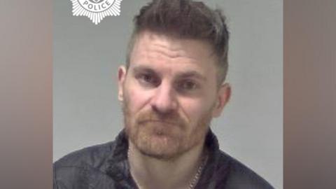 A man with spikey dark grey hair is pictured wearing a black coat in front of a grey/neutral background.