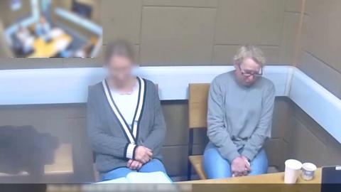 Two women are sat down in a room on wooden chairs. The woman on the right of the image has her face blurred and is wearing a grey cardigan with a white stripe on the sleeves and mid-section. The woman on the left is wearing a grey jumper and glasses. The room is cream coloured with square and rectangular shapes in the wall.