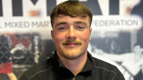 Ruairi O'Kane - a young man with brown hair and a mustache, wearing a black top 