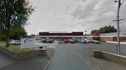 The Geoff Moulder Leisure Complex - a single-storey brick and metal building with a red roof. A car park with several cars is in front, along with a pedestrian crossing. A white sign with blue text marks the entrance. Trees and a cloudy sky are visible in the background.