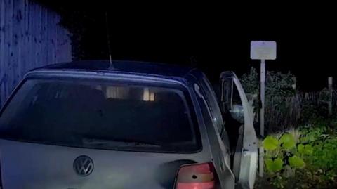 Photo taken from behind a stationary silver VW Golf car. The lights are off and the door to the drivers' side is open. There is a small faded street sign in front which may be indicating water nearby but it is too faded to see.