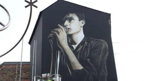 A mural of Ian Curtis on a side of a house in Macclesfield. He is holding a microphone to his lips and his left arm is visible as well as his wedding ring.  