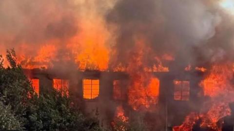 The Saxonvale fire - there are big orange flames and lots of thick, grey smoke. Through the flames, the remnants of the derelict building can be seen.