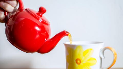 Tea being poured from a red teapot into a mug which is white, with a yellow flower on the front. All against a while background.