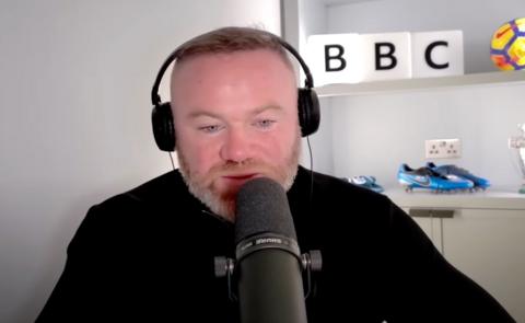 Wayne Rooney, wearing headphones and a black top, speaks into a mic