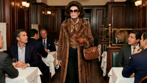 AI generated image by Gucci showing an AI lady wearing a glamourous fur coat and sunglasses walking through a wood panelled room. There are with dining tables with white table cloths and male guests in suits who are all looking at the lady.